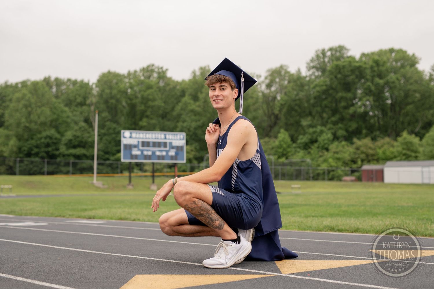A graduate in navy blue attire kneels on an athletic track with bleachers and trees in the background.