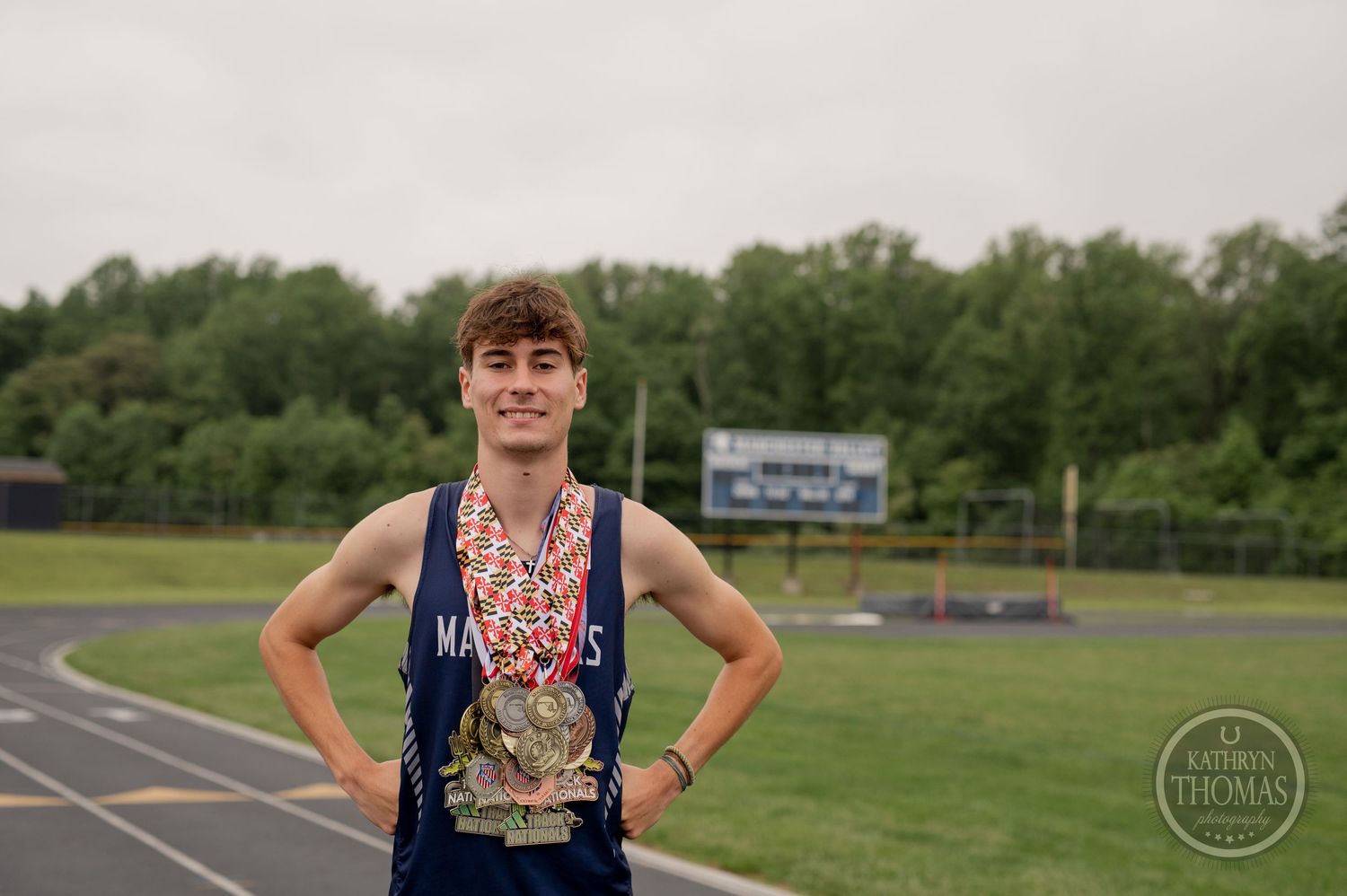 An athlete stands with hands on hips wearing medals on an outdoor track field.
