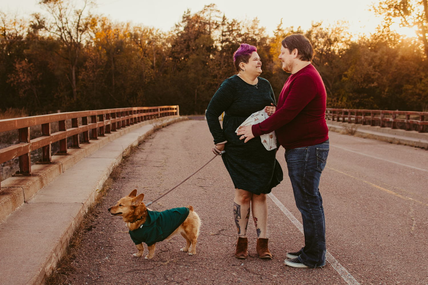 Couple and their small dog in winter clothes share a moment on a wooden bridge at sunset.