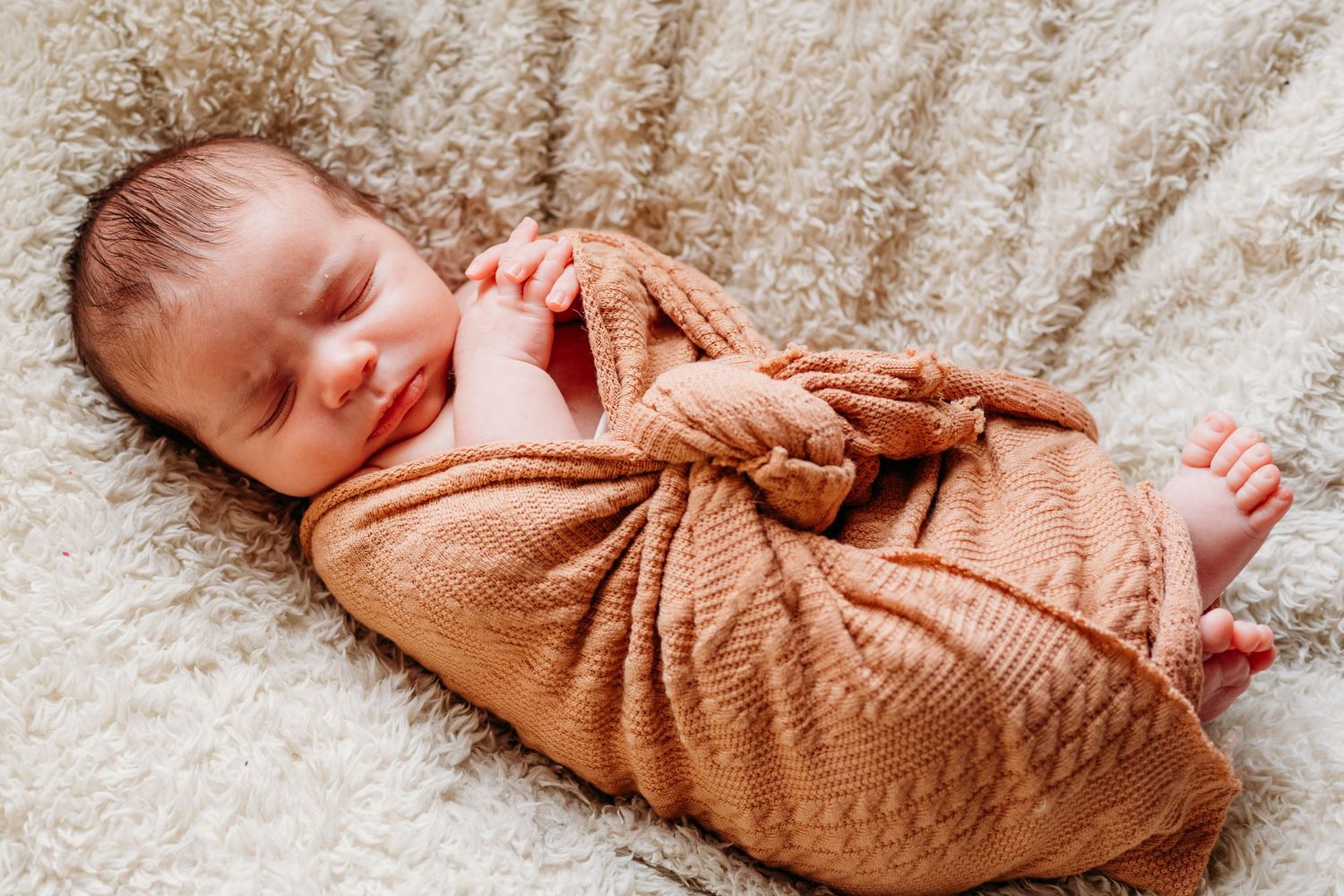 Newborn baby wrapped in soft brown swaddle blanket lies peacefully on textured beige surface.