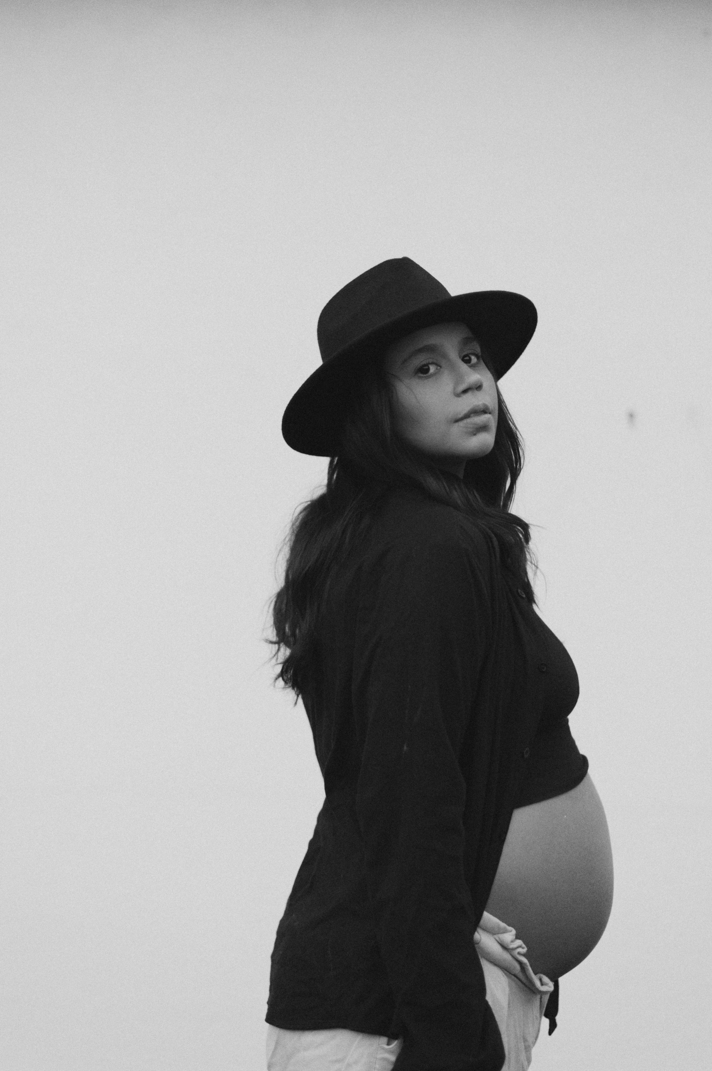 Stylish maternity portrait with woman wearing black hat against minimal background.