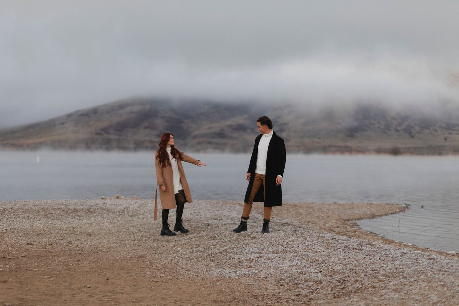 Couple walks along the shoreline with foggy mountains behind them.