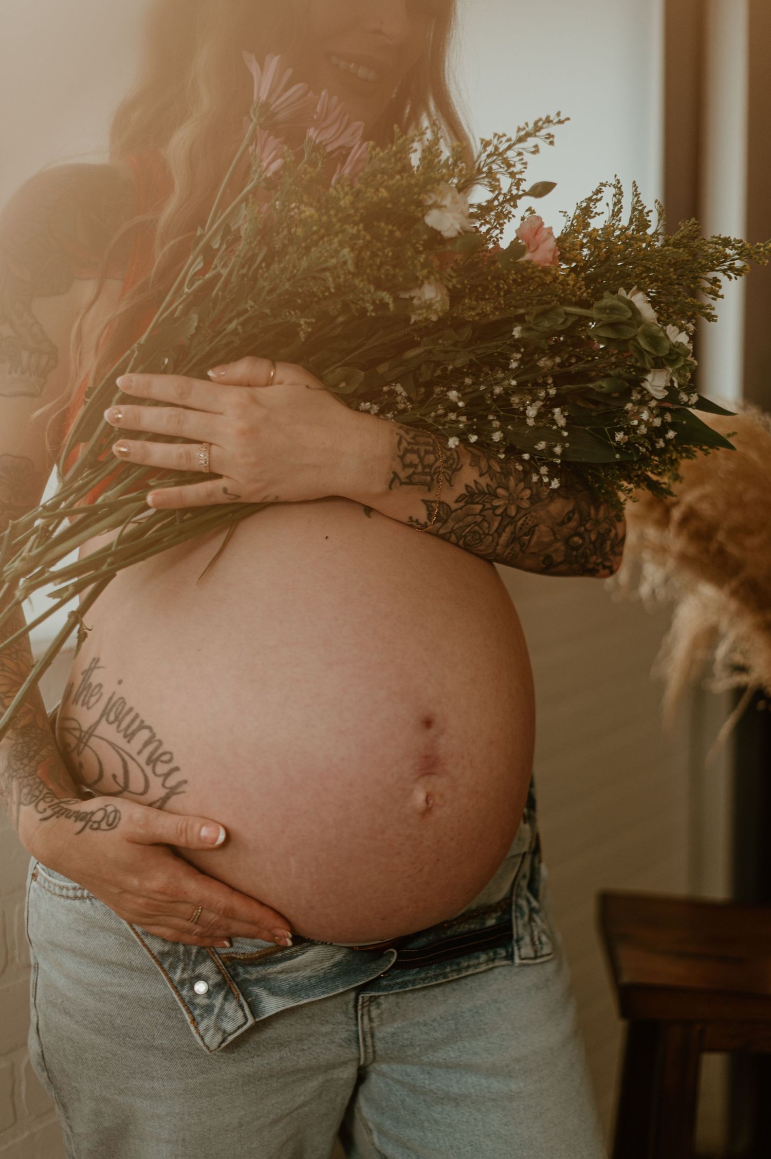 Close-up of pregnant belly with dried flower bouquet in warm sunlight.