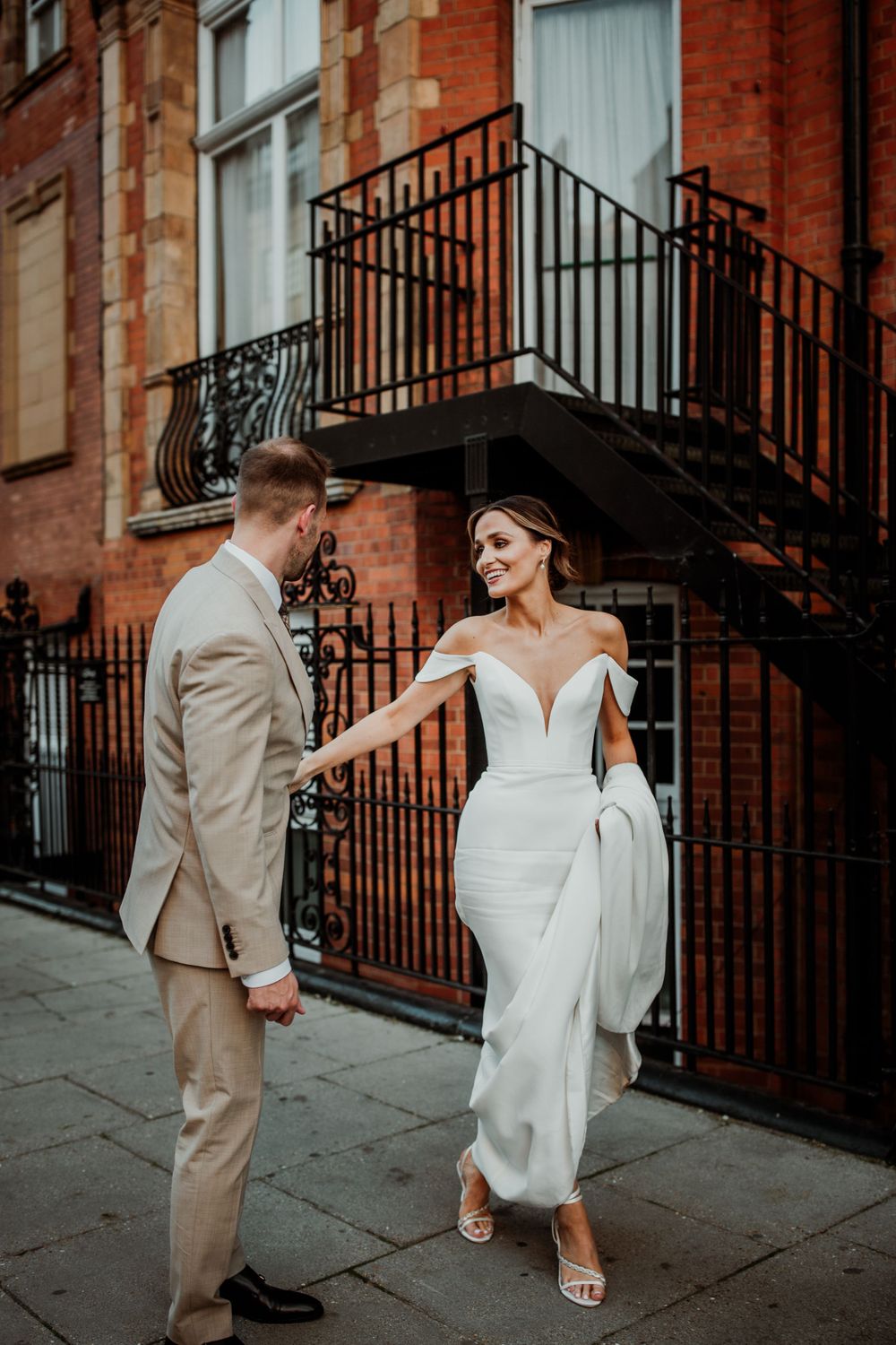 A wedding couple in elegant attire pose on a city street by a historic red brick building with iron railings.