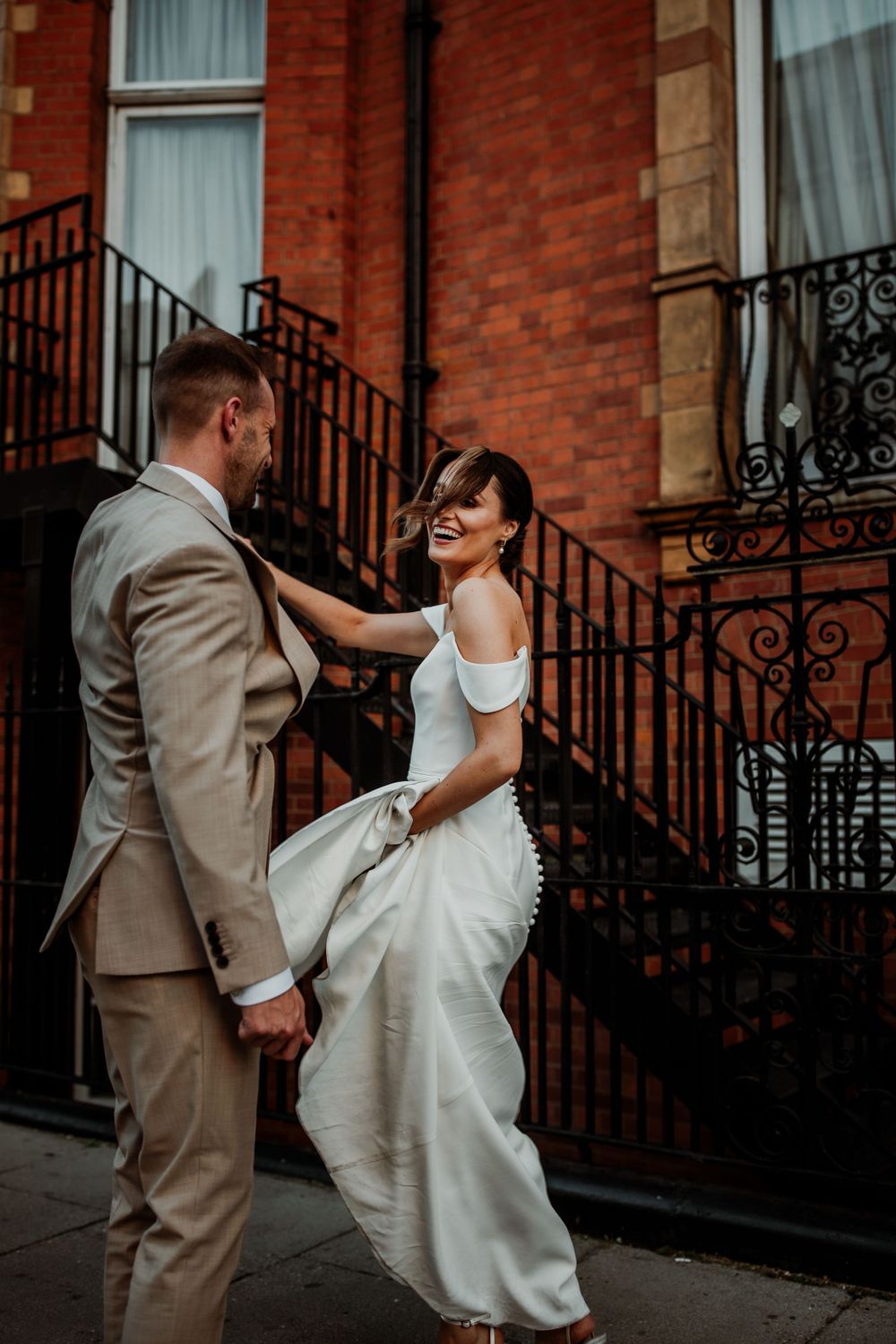 Two people share a joyful moment in wedding attire outside a historic brick building with ornate iron railings.