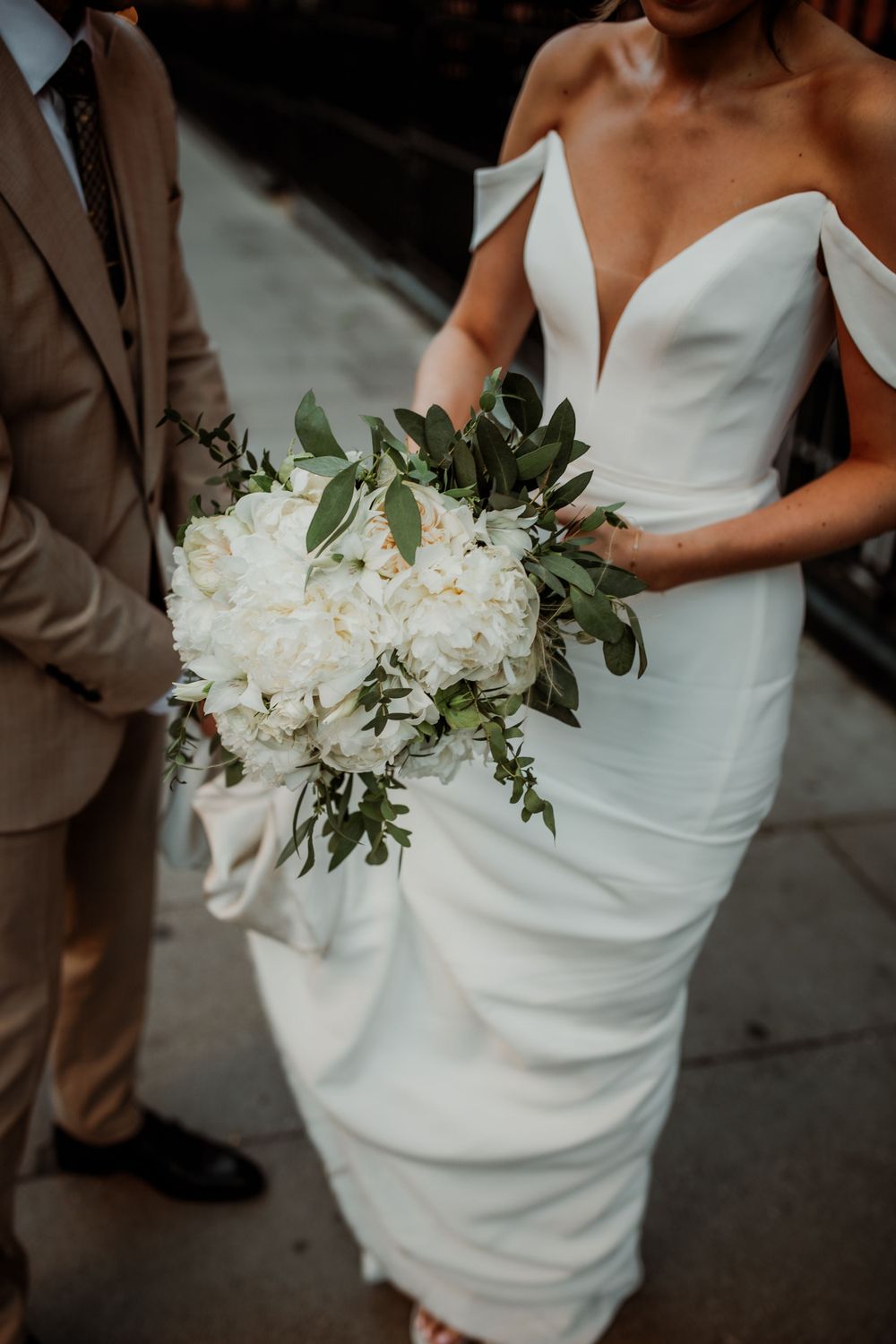 A stunning white off-shoulder wedding dress with an elegant white peony bouquet accented with fresh greenery.