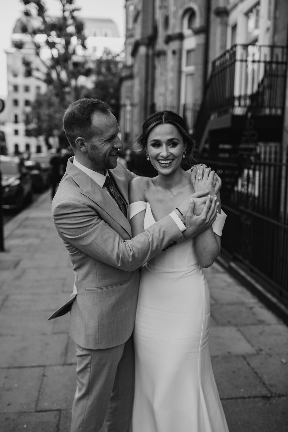 Black and white wedding photo of couple on city sidewalk, bride in sleek gown and groom in light suit.