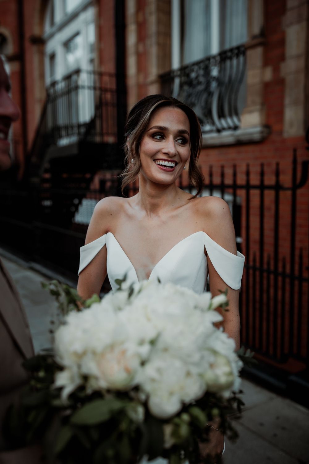 A bride wearing an elegant off-shoulder white wedding dress holds a stunning bouquet of white flowers on a city street.