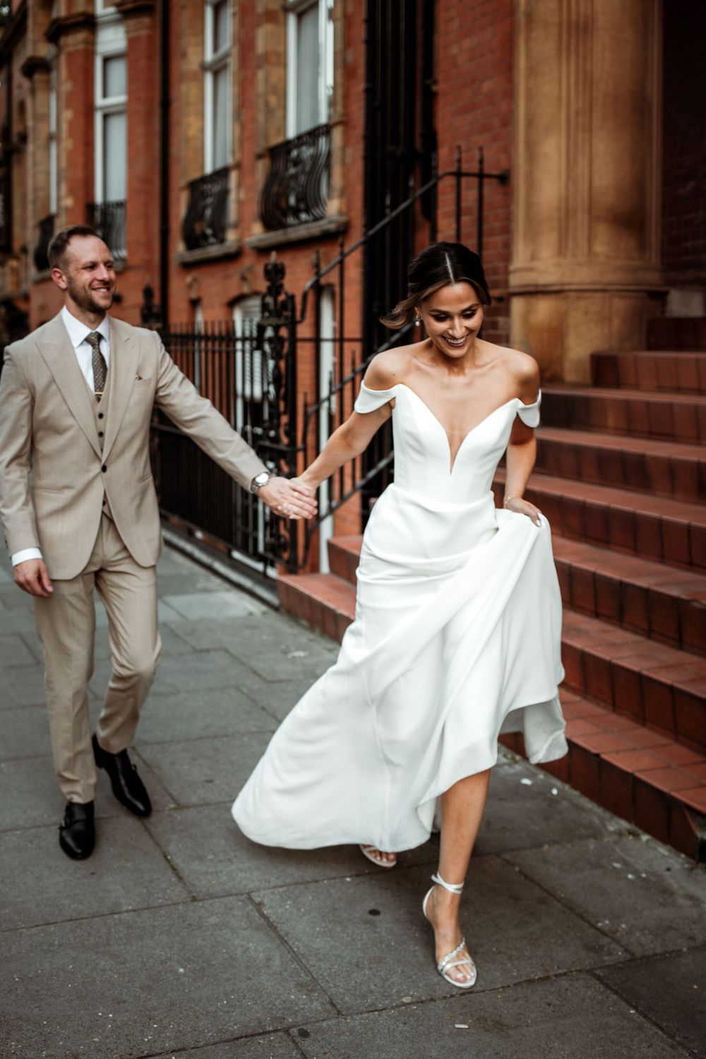 A couple in wedding attire descends red brick steps, with the bride in an off-shoulder white gown and groom in light beige suit.