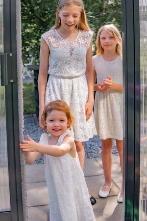 Three children in white lace dresses standing by a glass doorway on a sunny day.