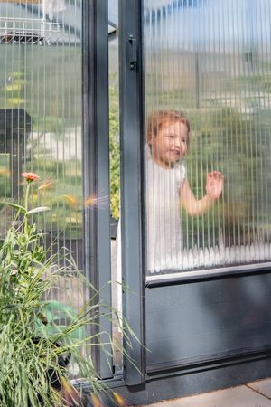 A child peeking through a glass door with plants visible in the background.