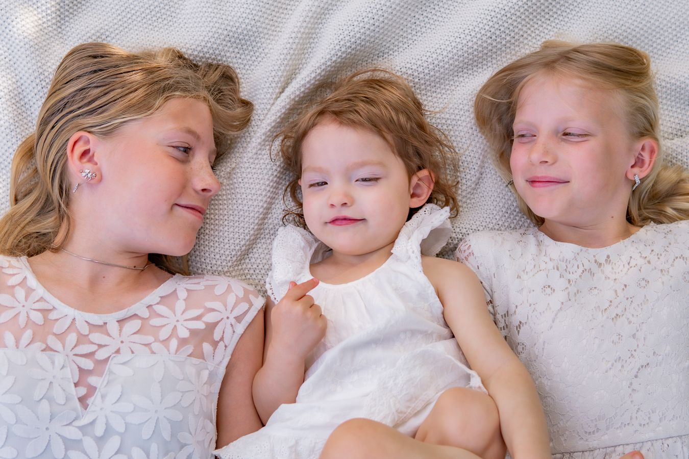 Three children in white lace dresses resting together on soft fabric.