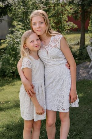Two children in matching white lace dresses standing together in sunny garden.