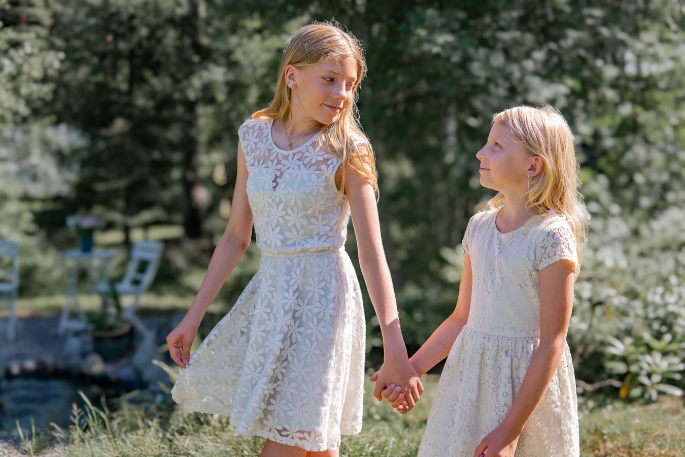 Two girls in white lace dresses hold hands while walking outdoors on a sunny summer day.