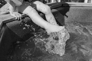 Black and white image showing splashing water and legs at fountain edge.
