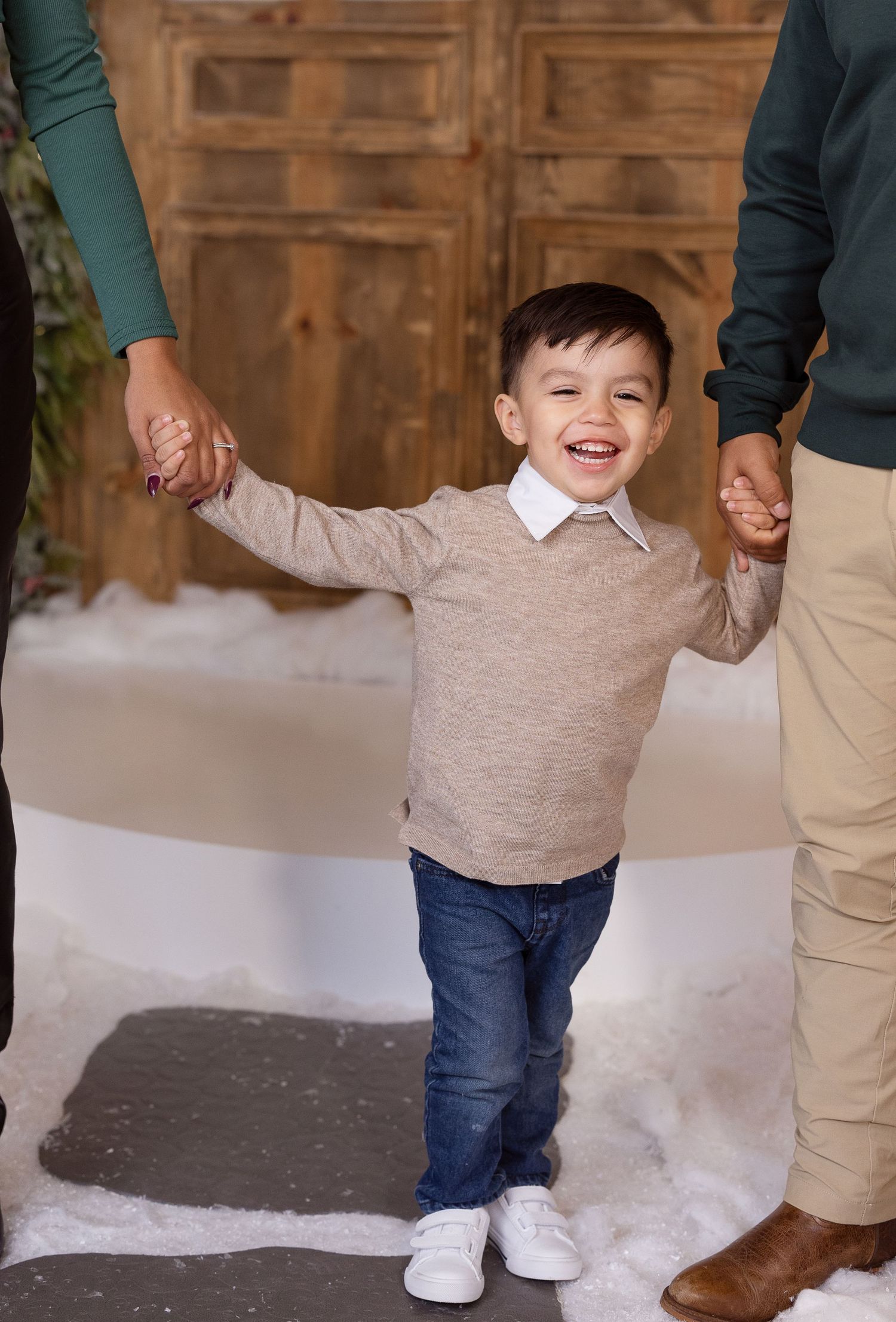 A small child in a beige sweater and jeans holds hands with others while standing in snow against a wooden door.