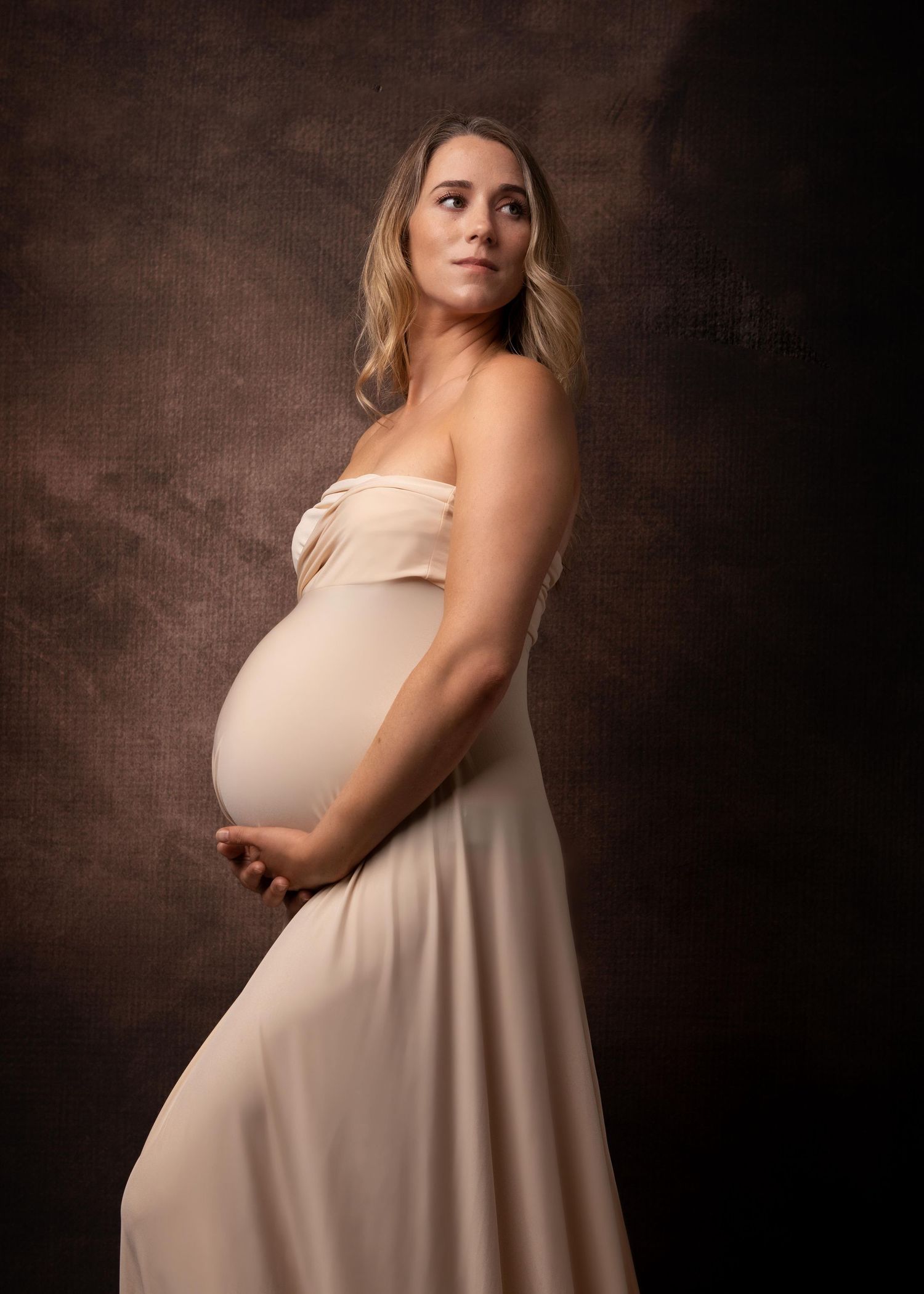 A pregnant woman in a flowing peach strapless dress poses against a dark brown textured backdrop during a maternity photoshoot.