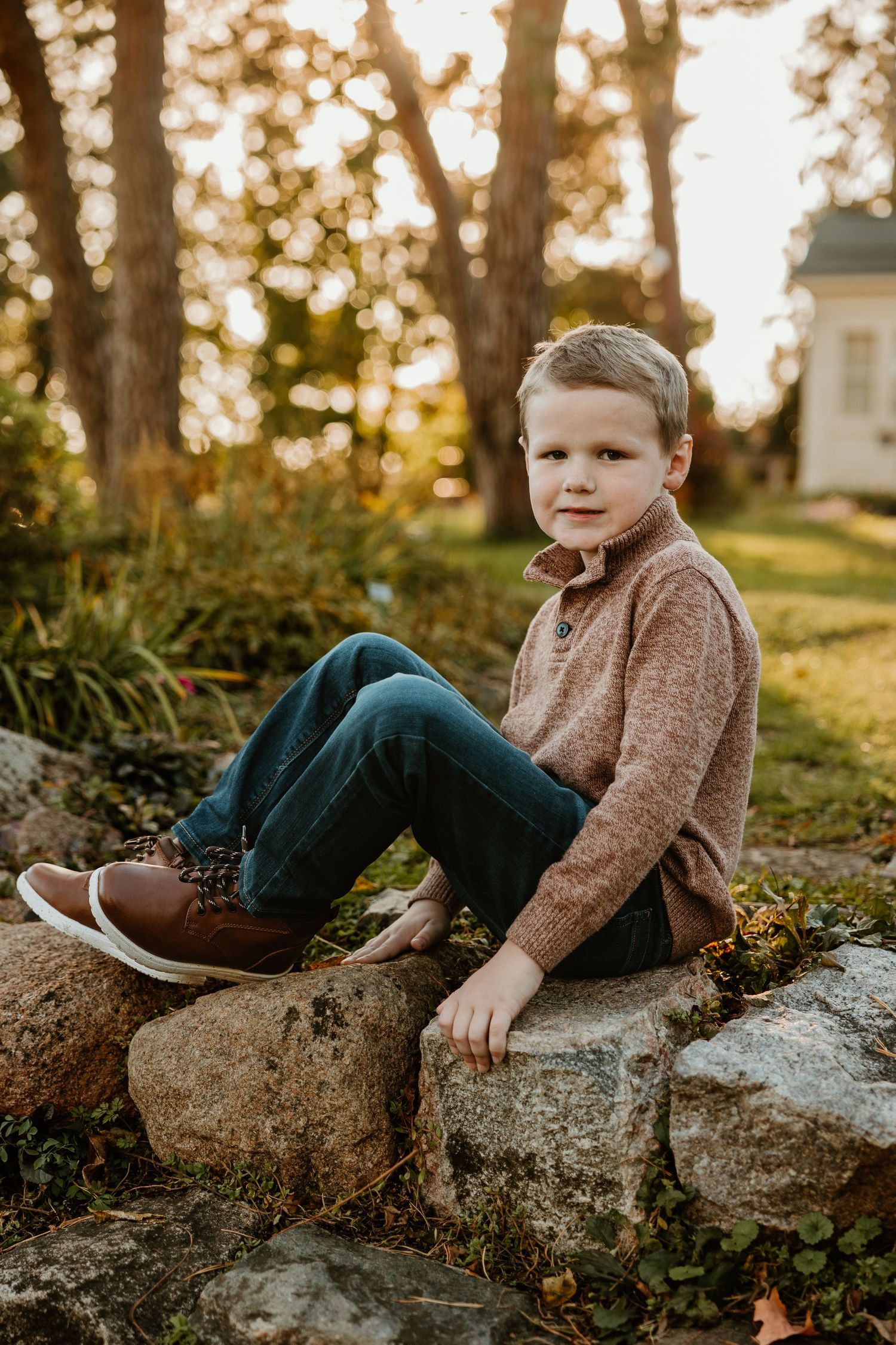 Child sits on large rock wearing brown sweater during golden hour in autumn garden setting. Michigan Metro Detroit Novi Northville Livonia