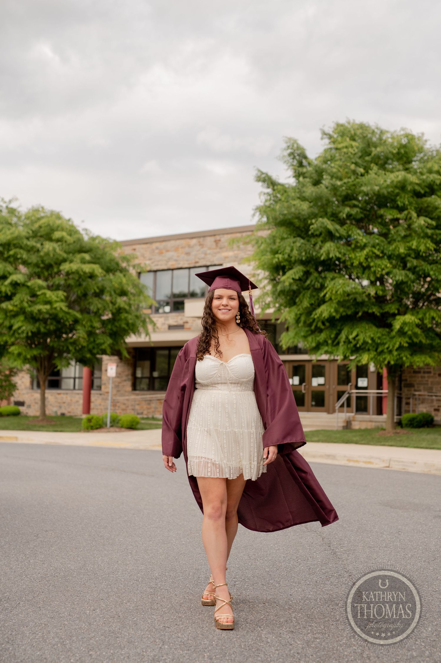 Graduate walks confidently in burgundy graduation attire and white dress outside high school.