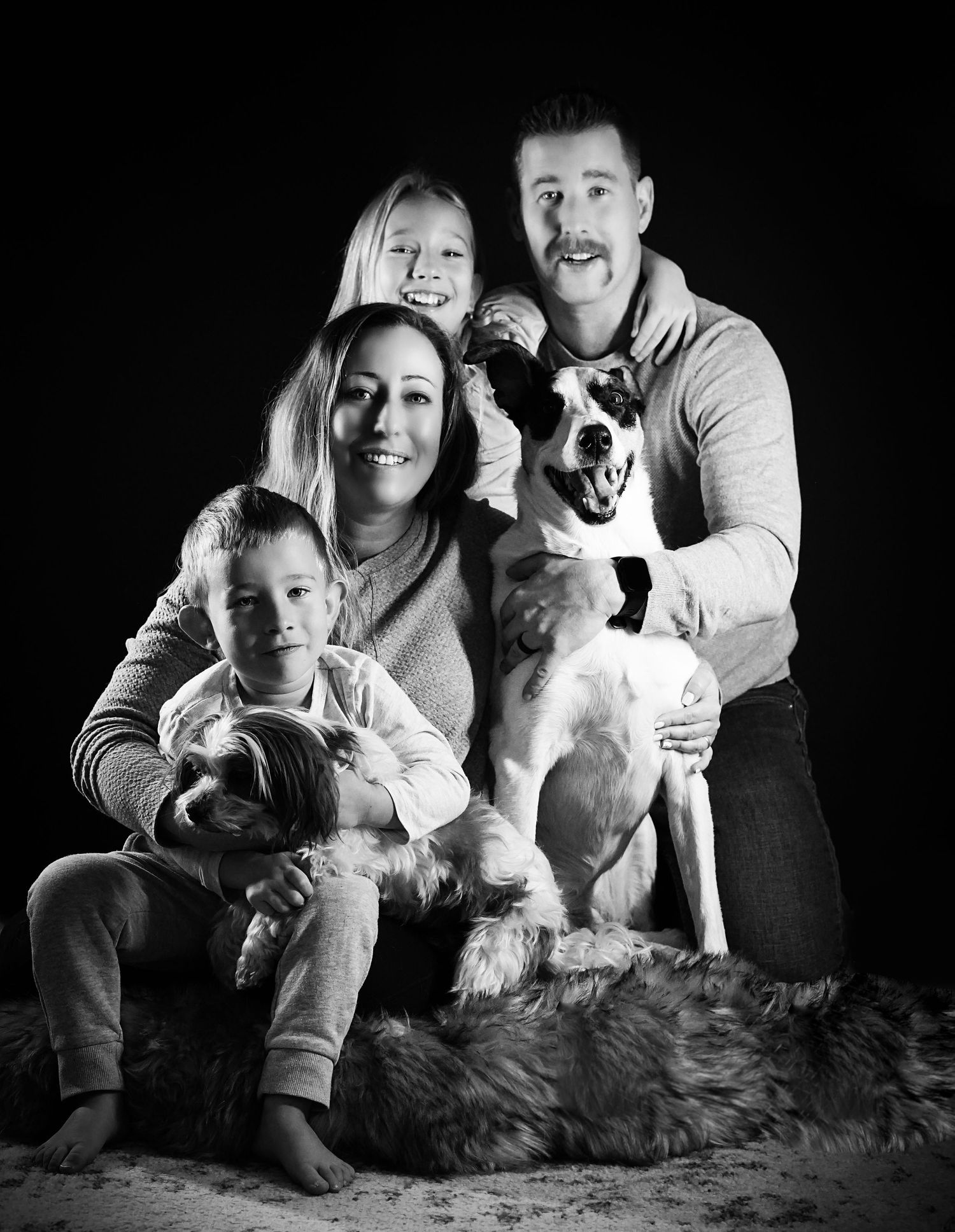 Black and white studio portrait of a family with two dogs on a dark background.