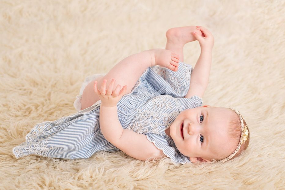 six month baby girl in blue dress on back and grabs her toes while looking at the camera in studio milestone session, Baltimore Ontario*