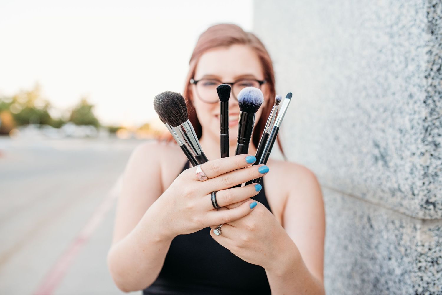 Woman holding a fan of professional makeup brushes in front of her face outdoors.