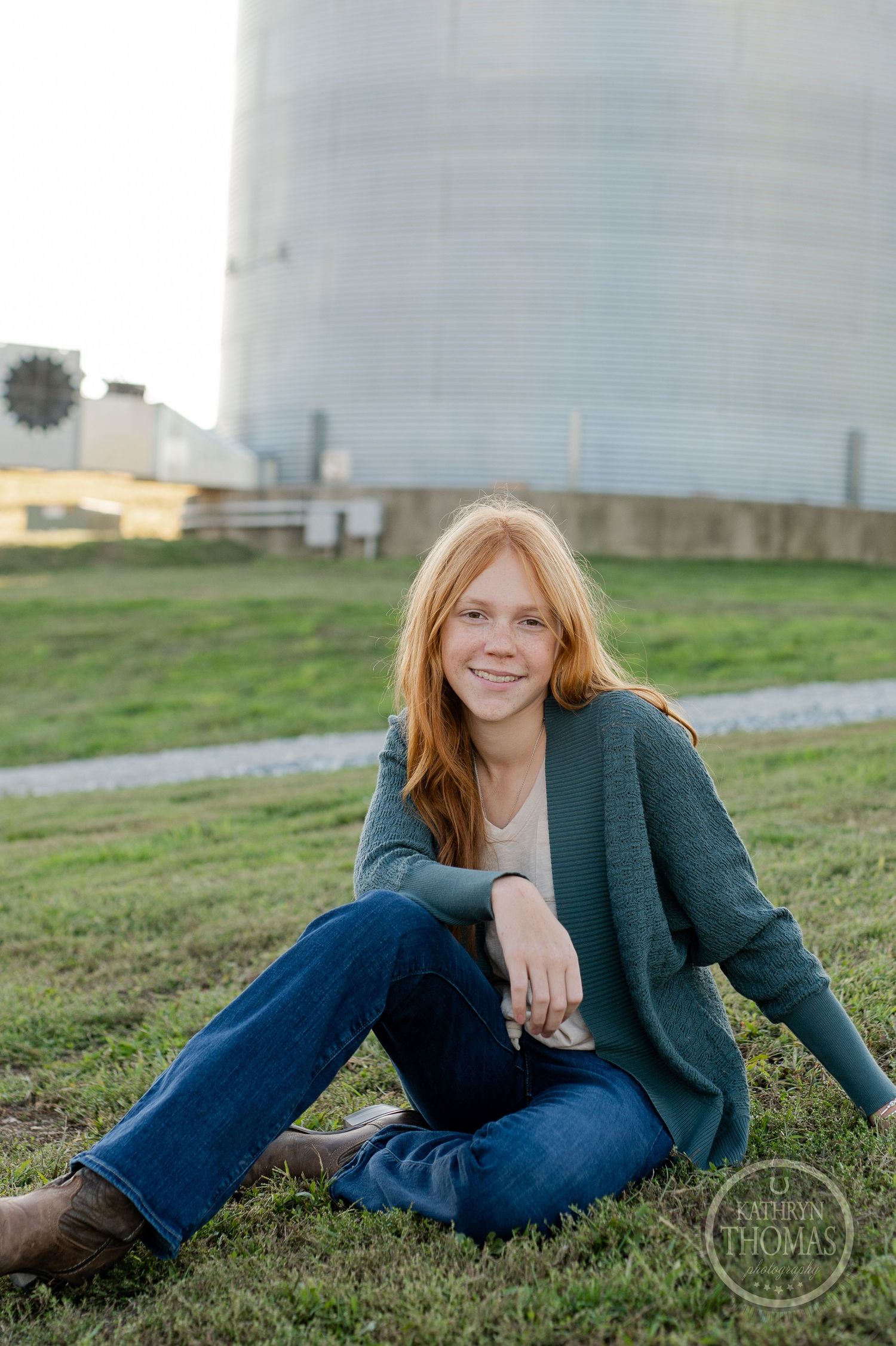 Someone in a green cardigan and jeans relaxes on grassy hill with large grain silo in background.