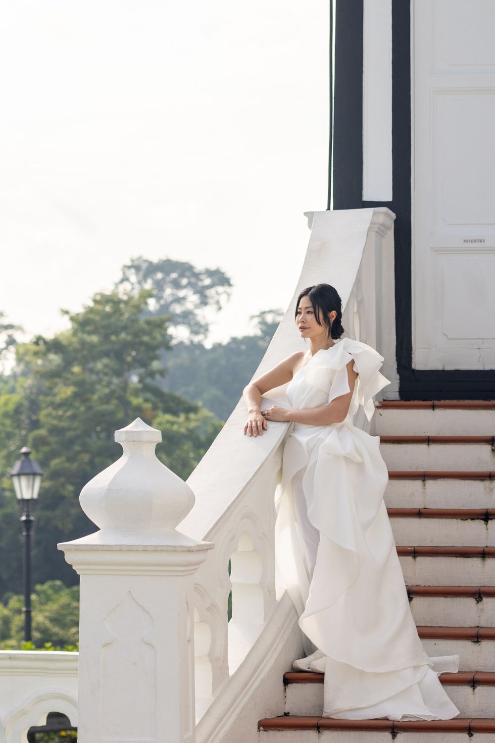 Bride in an elegant white wedding dress poses gracefully on white marble stairs of a colonial-style building.