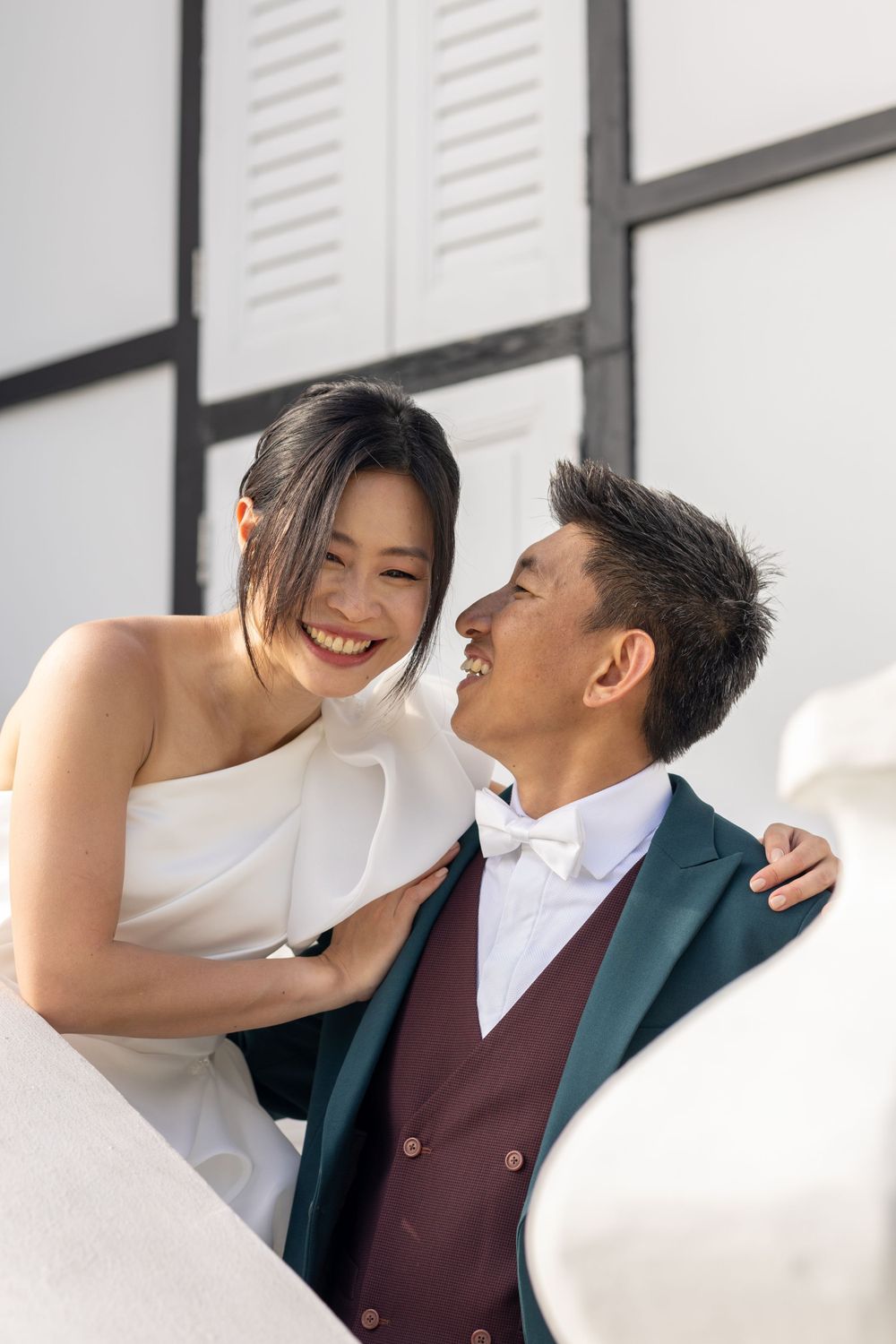 Happy couple sharing joyful laughter during their wedding photoshoot against white building.