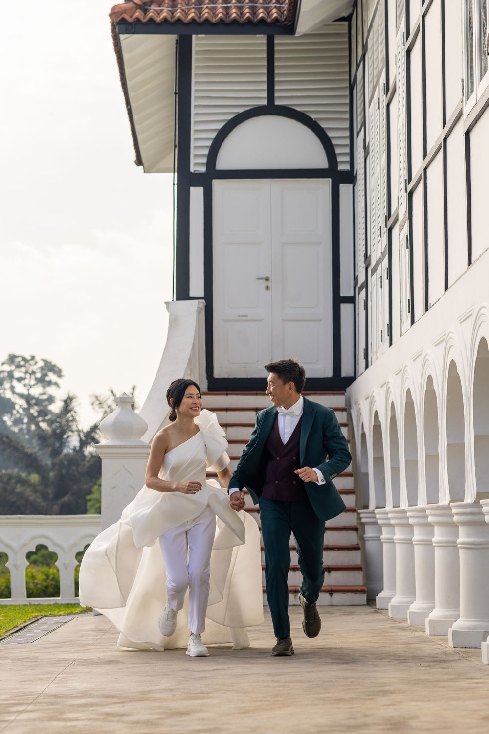 Playful newlyweds running along colonnade in wedding attire and sneakers.