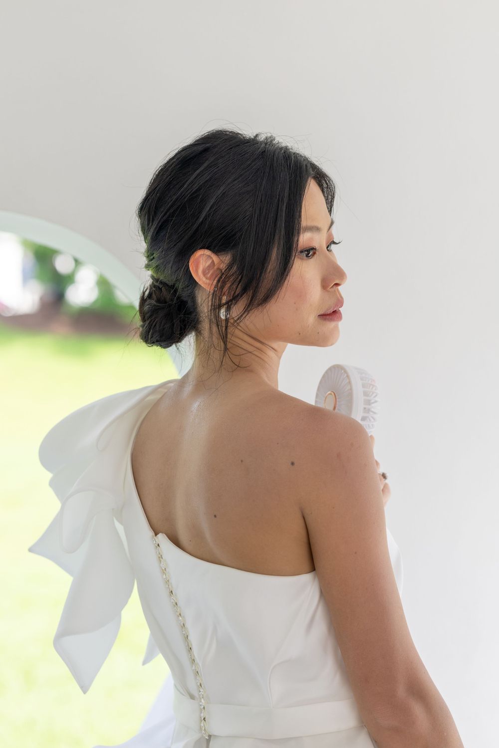 A bride in a white one-shoulder dress with a bow detail gazes sideways near a window in soft natural light.