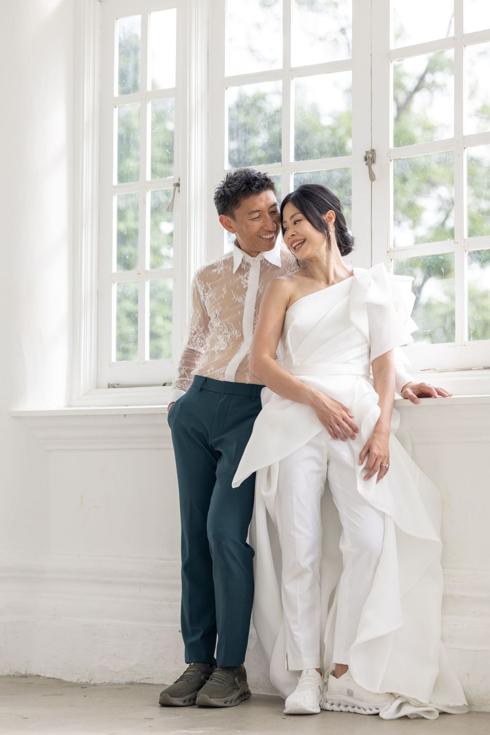 A couple poses romantically by large windows in wedding attire, with the bride wearing a modern white jumpsuit.
