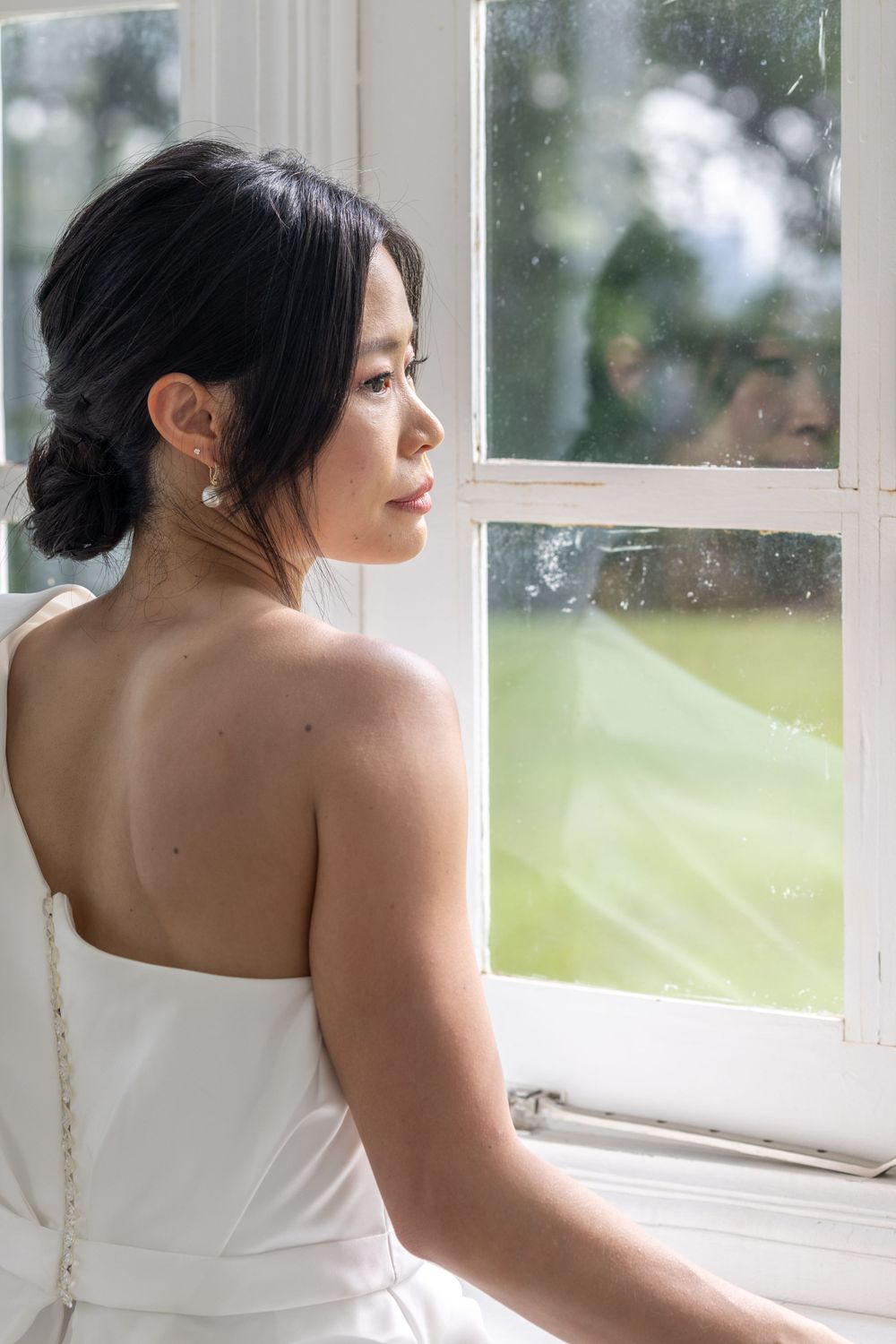 A bride in a strapless white dress gazes thoughtfully out a window in natural light.