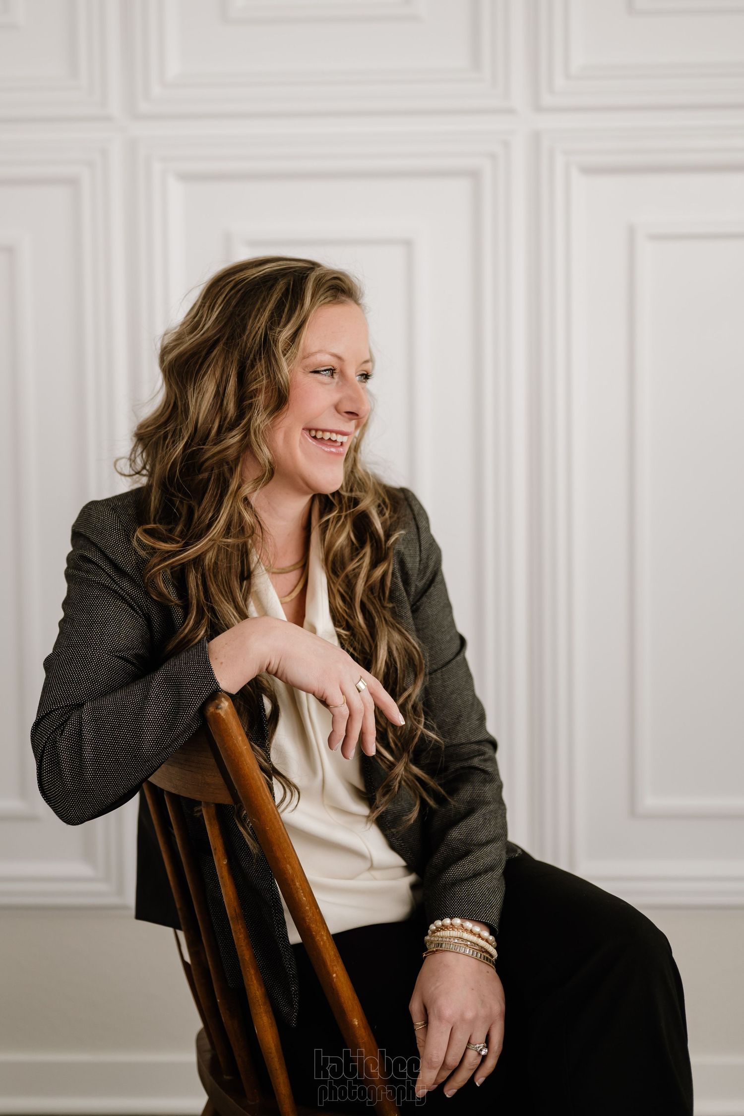 A smiling figure in a dark blazer and cream top sits casually against a chair in front of a white paneled wall.