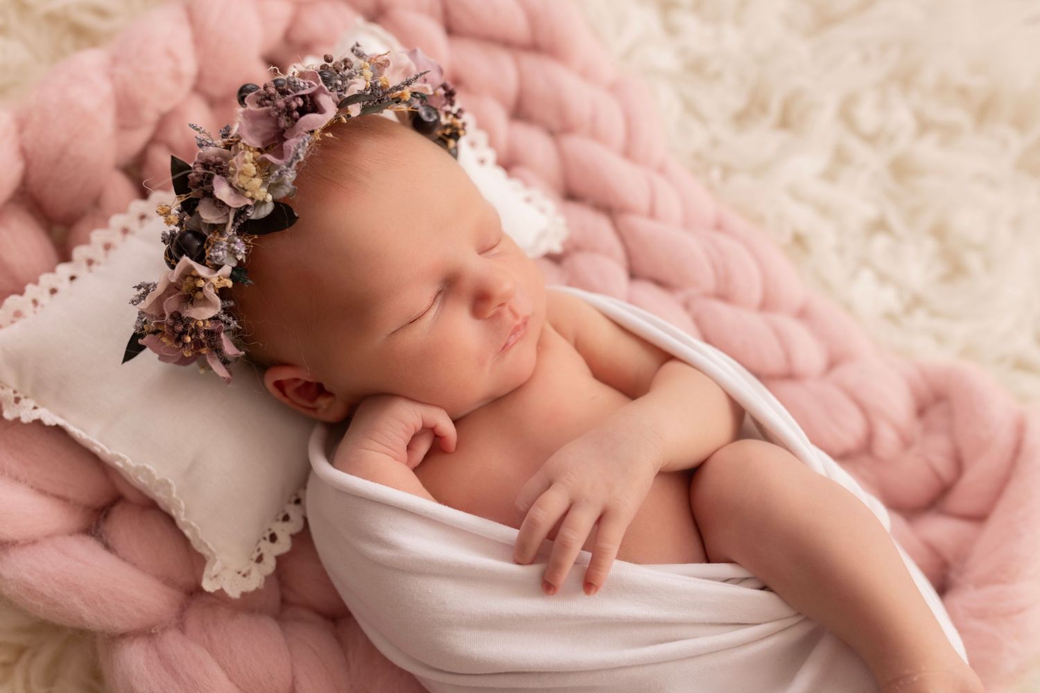 A peaceful sleeping newborn wearing a floral crown lies on soft pink bedding with a small white pillow.