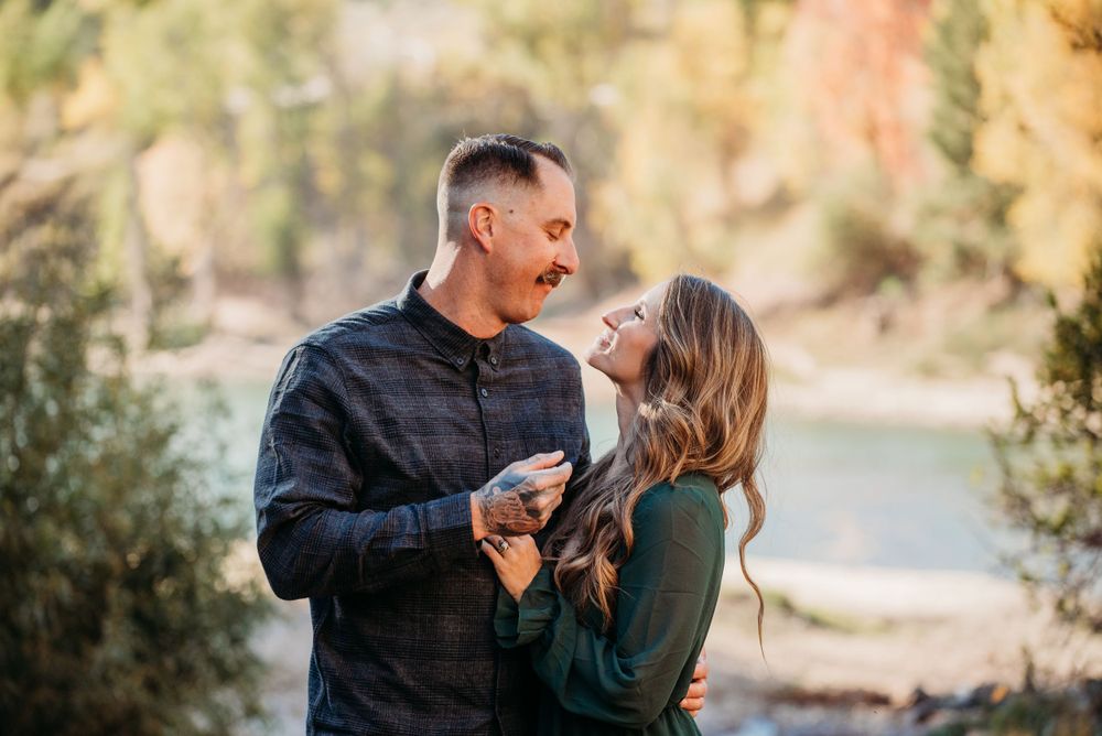 Happy couple laughing and embracing outdoors with golden autumn trees and a river behind them.