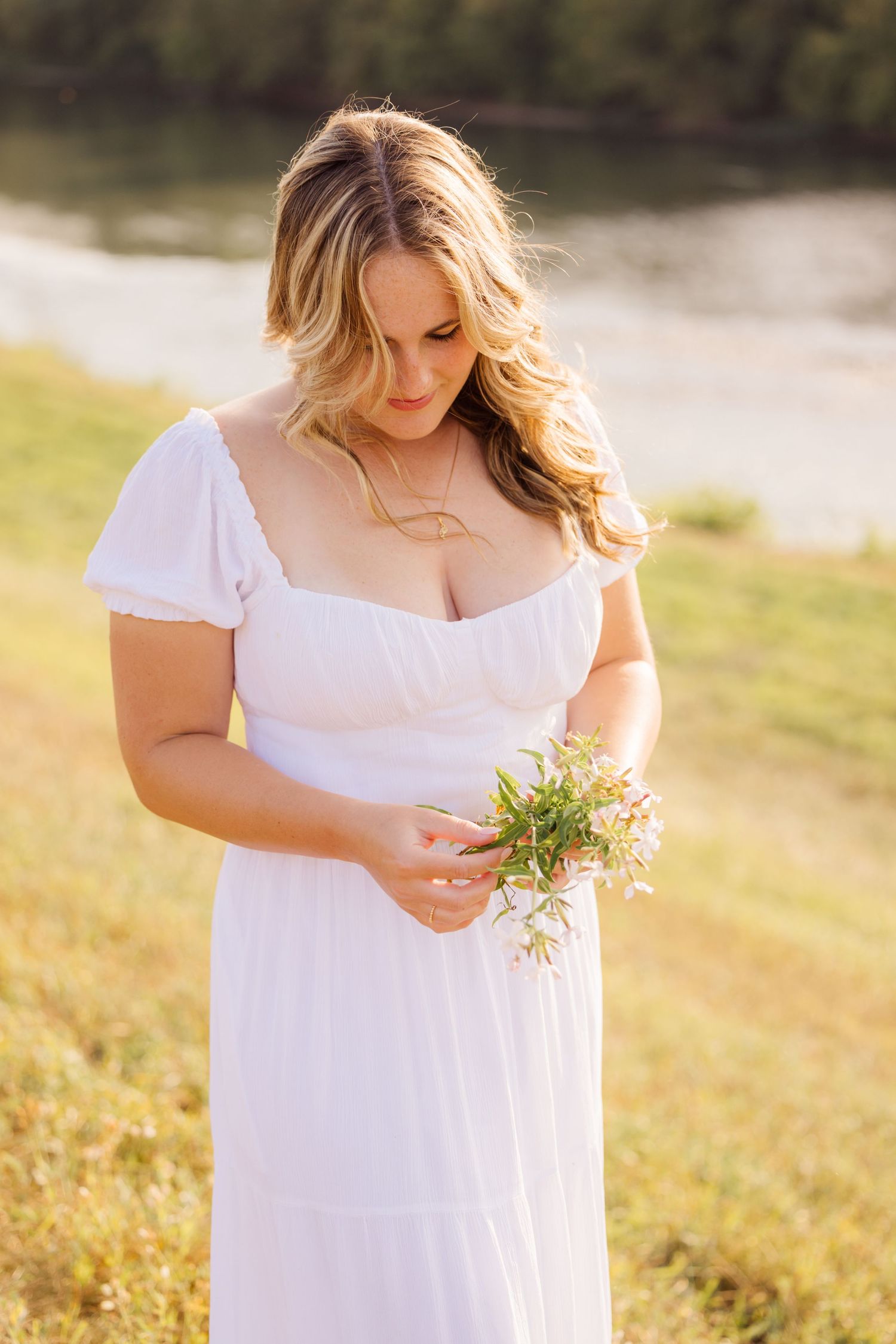 Woman in white summer dress holds small wildflowers while standing in sunlit field near water.