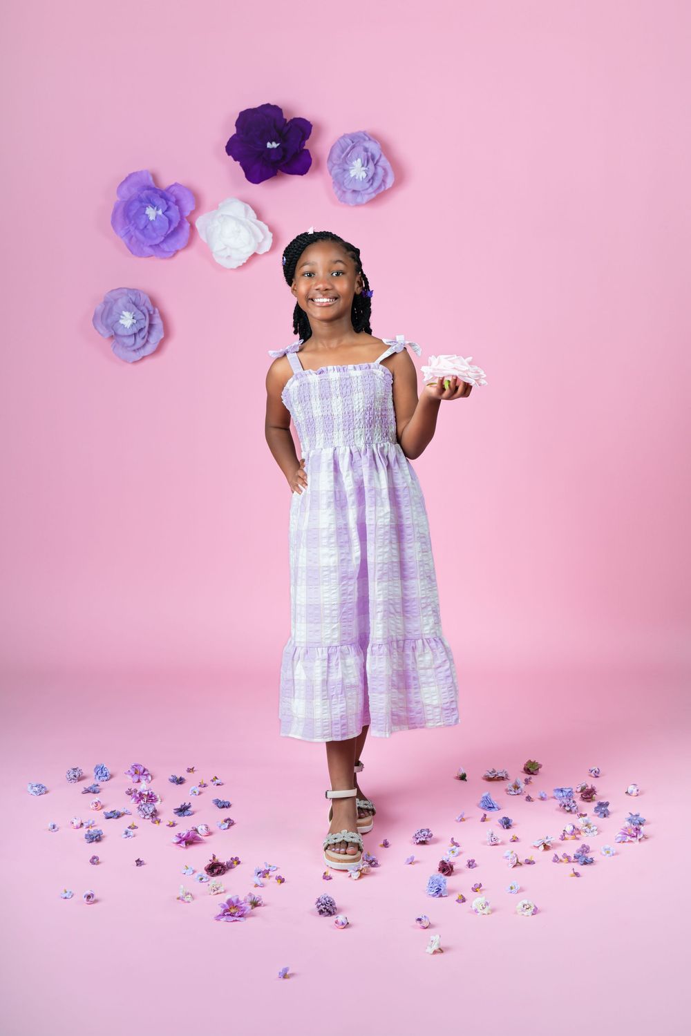 Child in white summer dress posing with purple flowers on pink background.