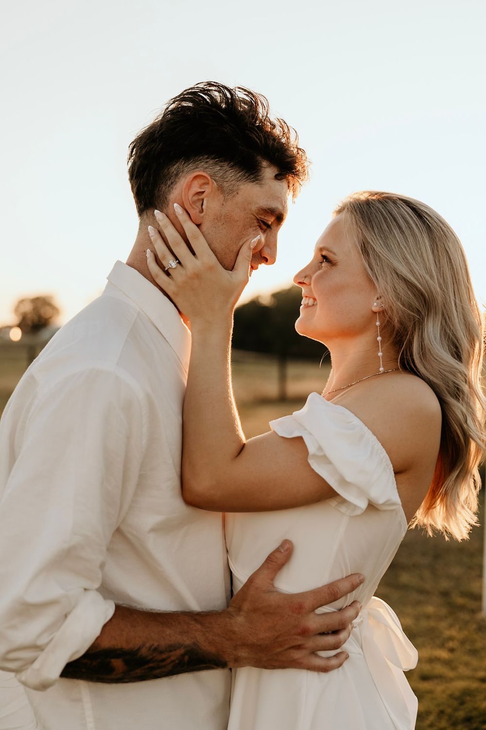 Romantic couple in white outfits embracing during golden hour in a rural field setting.