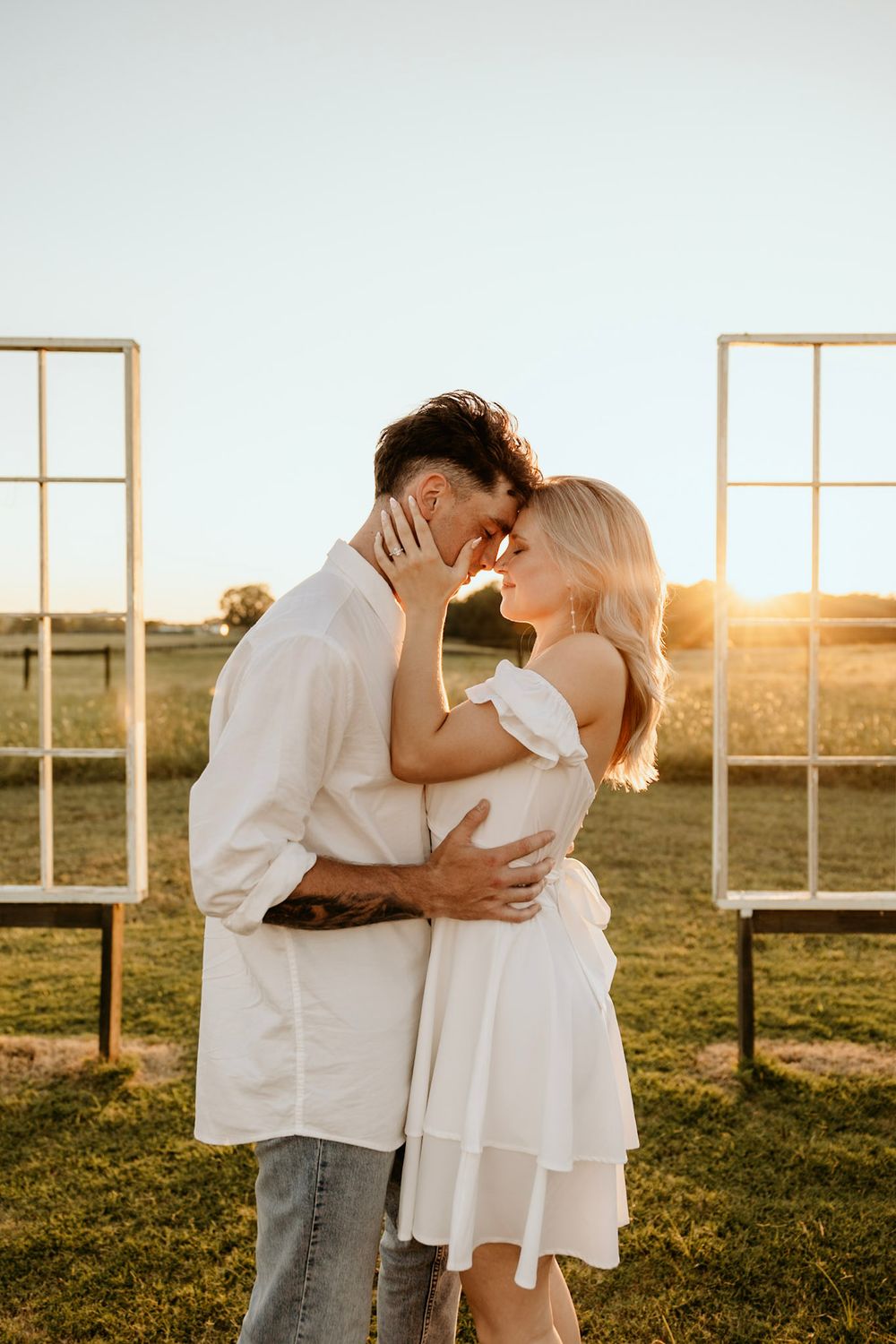 Romantic couple embraces in white outfits against golden sunset with window frames in outdoor setting.