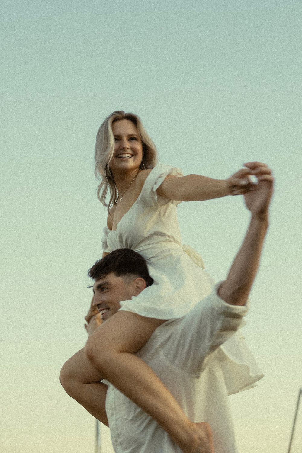 A couple in white clothing playfully interacts outdoors during sunset with one person carrying the other on shoulders.