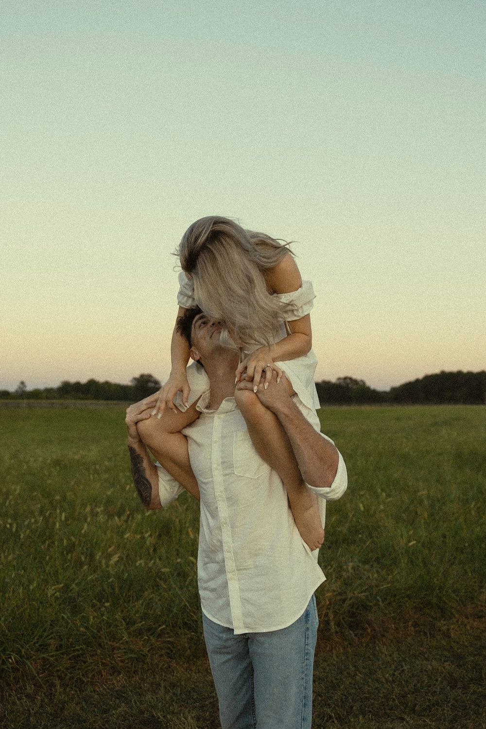 A couple sharing a playful moment in a grassy field at sunset as one lifts the other up from behind.