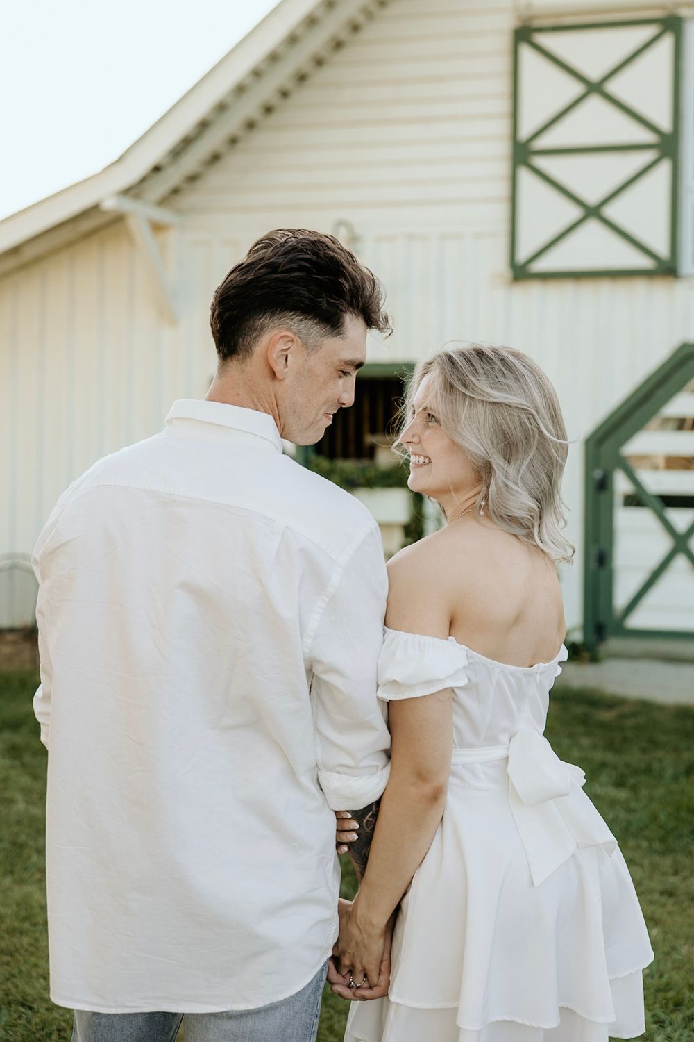 A romantic couple in white outfits hold hands while standing in front of a rustic barn with green accents.