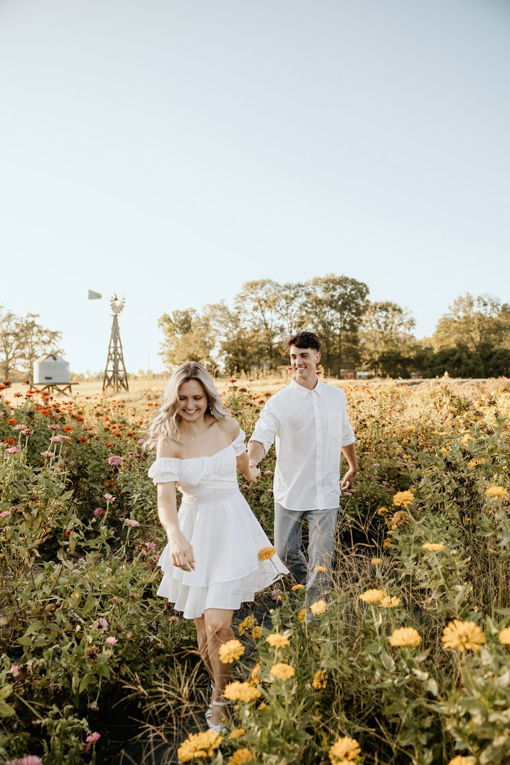 A couple walks through a wildflower field with a rustic windmill in the background during sunset.