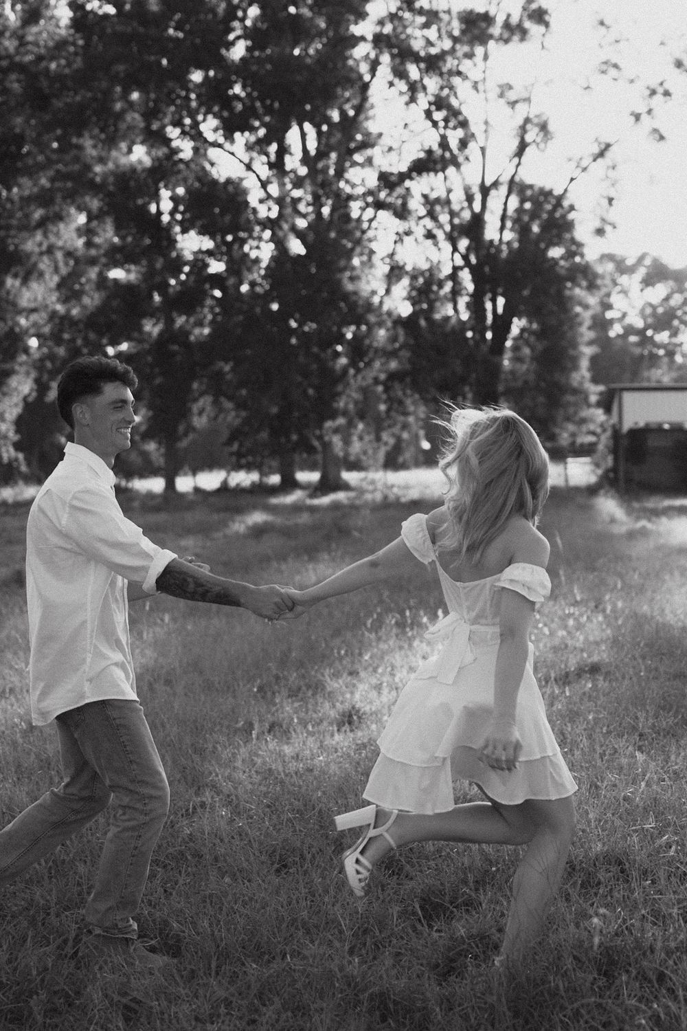 A couple in casual attire holds hands while running through a sunny field in black and white photography.