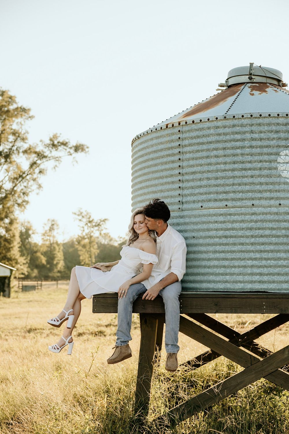 A couple shares a romantic moment sitting together on wooden support beams beneath a large grain silo at sunset.