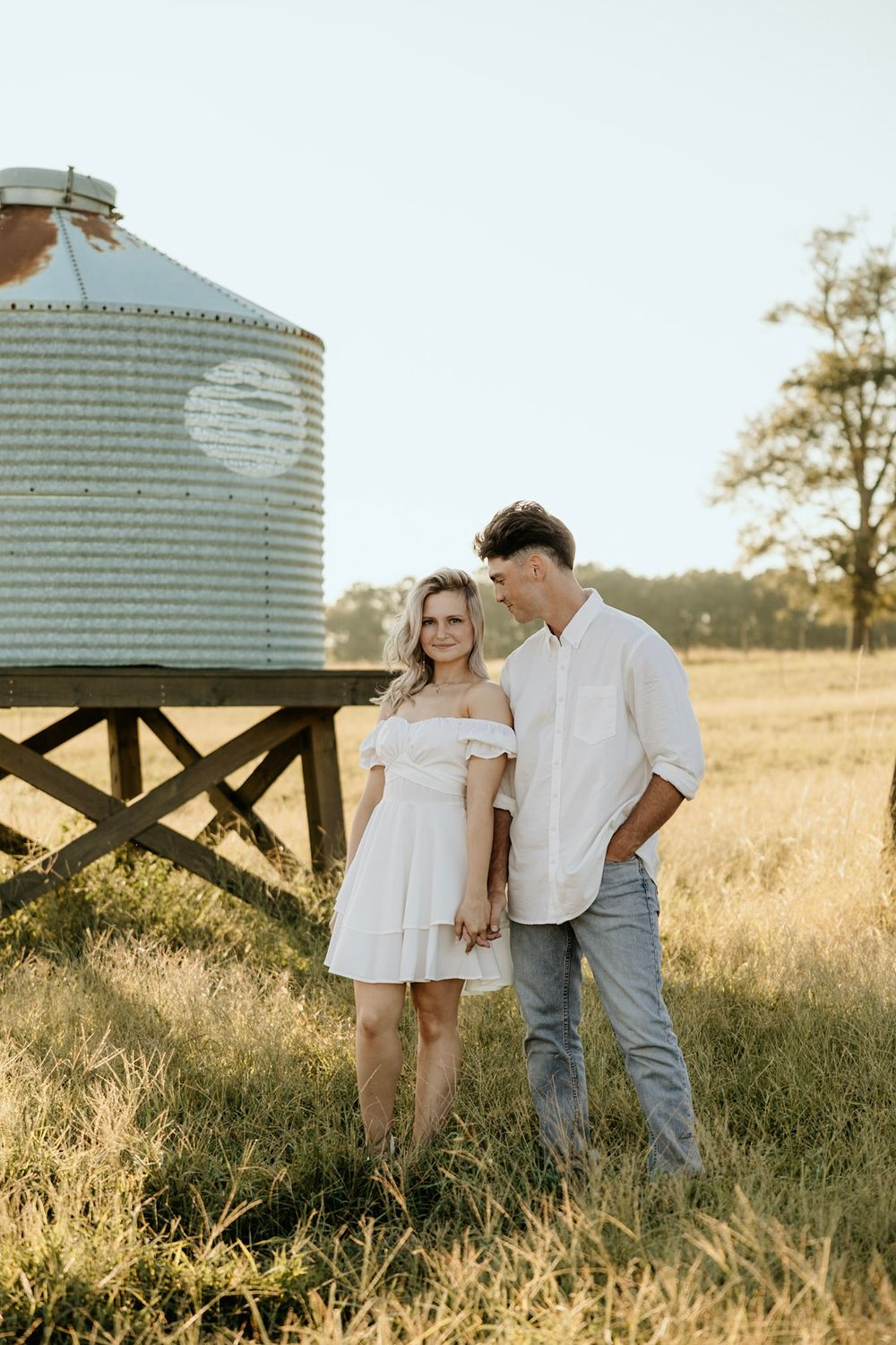 A couple stands together in a golden field at sunset next to a rustic grain silo in the countryside.