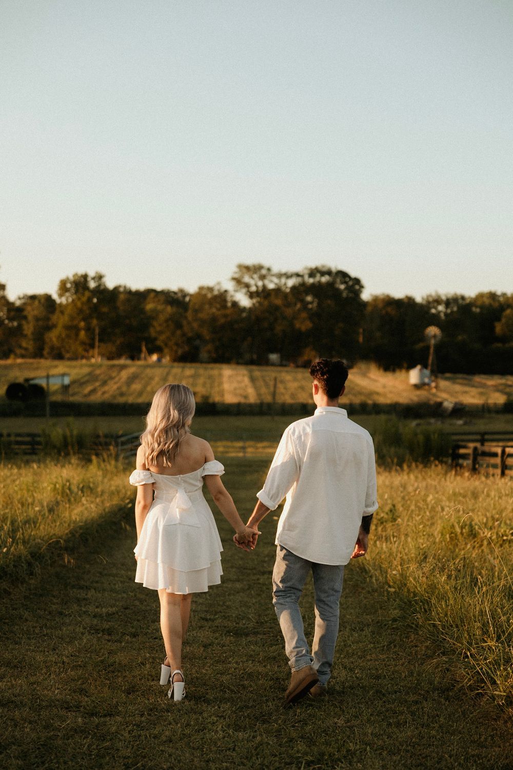 A couple walking hand in hand through a golden field at sunset wearing white and casual attire.