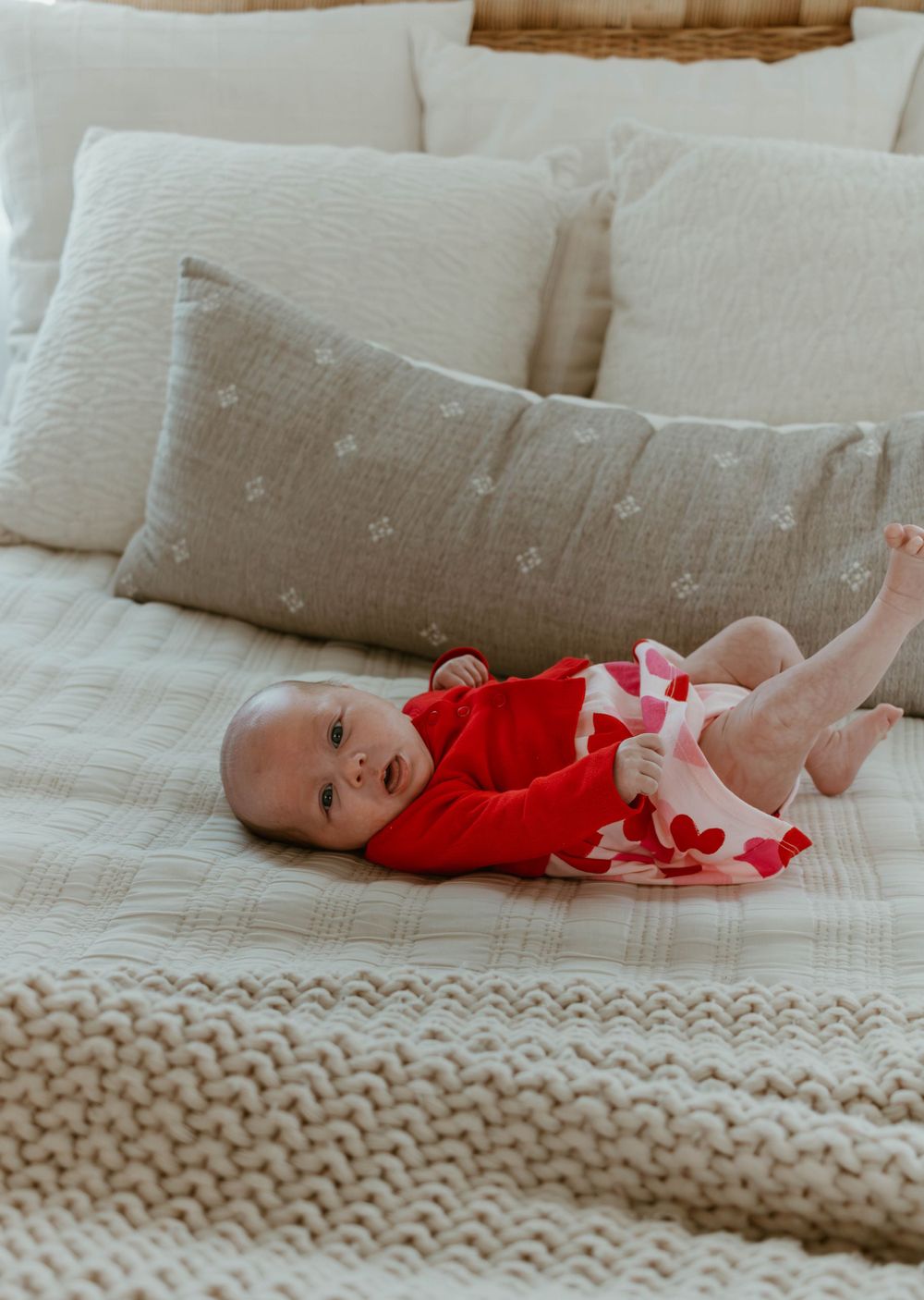 A small infant wearing a red outfit lies on a cozy bed with light colored pillows and a knit blanket.