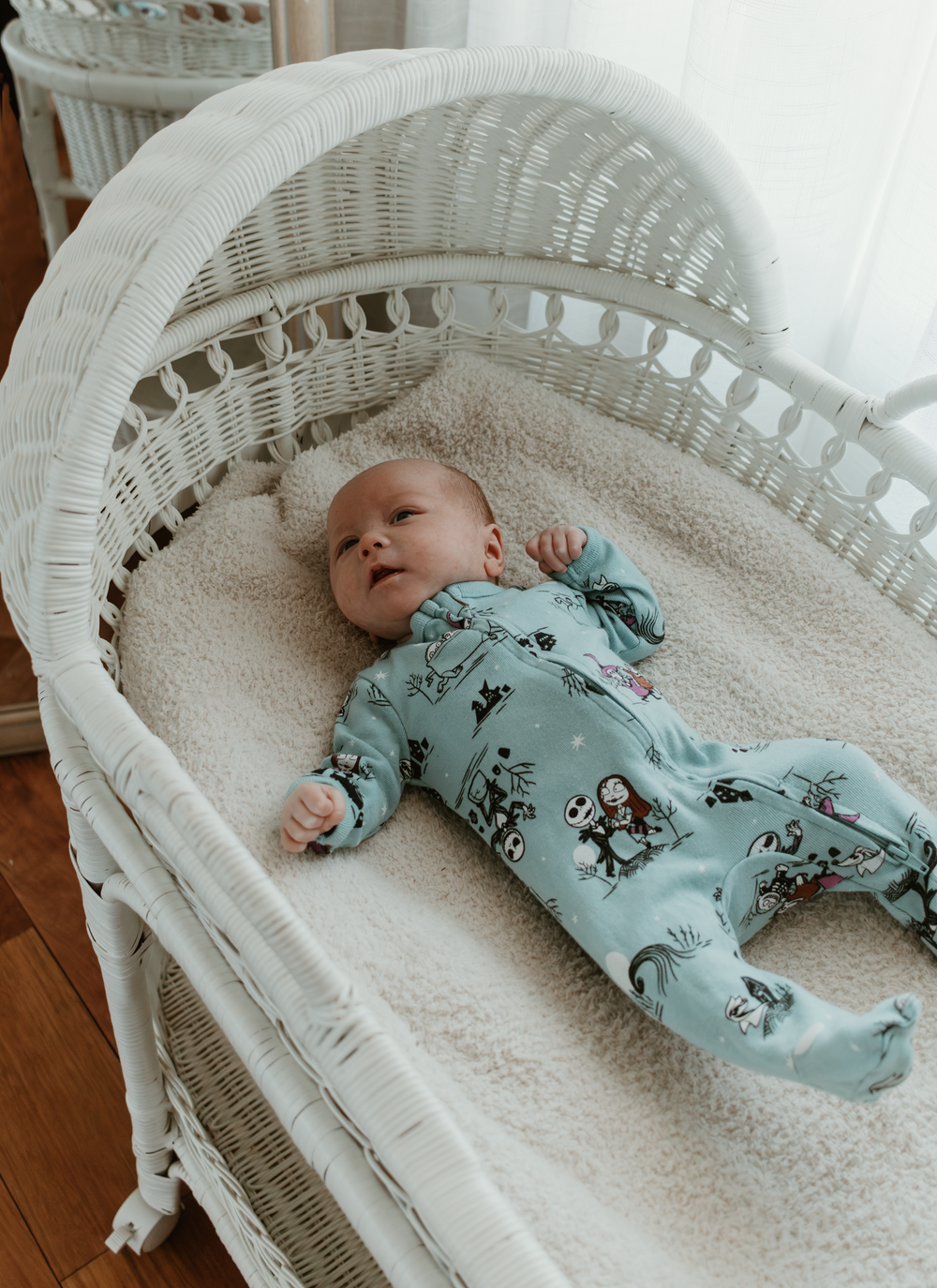 Baby in light blue sleeper resting peacefully in white wicker bassinet on soft bedding.